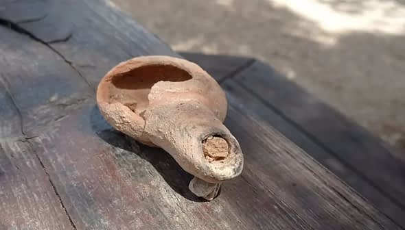 An ancient clay olive oil lamp with a spout and a round body resting on a wooden surface. - Olive Oil Times