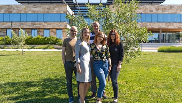 Five individuals standing together outdoors in front of a modern building with greenery. - Olive Oil Times