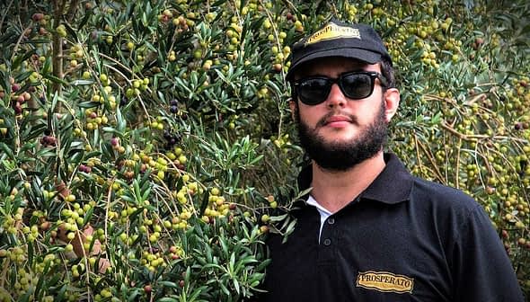 Man wearing sunglasses and a black cap standing next to an olive tree with green olives. - Olive Oil Times