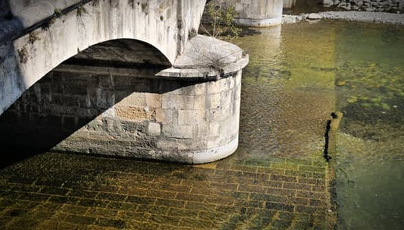 Close-up view of a stone bridge arching over a calm body of water with visible textures and reflections. - Olive Oil Times