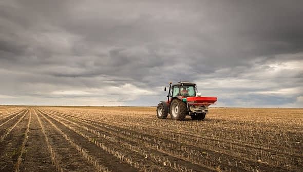 Red tractor equipped with a fertilizer spreader working in a field with rows of crops. - Olive Oil Times