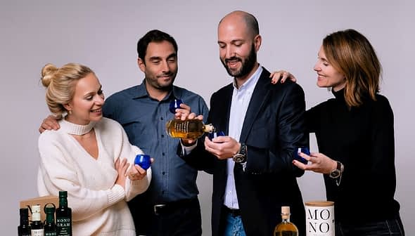 Four individuals holding olive oil bottles and small blue cups in a neutral setting. - Olive Oil Times