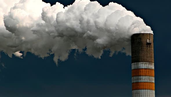 Tall industrial smokestack emitting large clouds of white smoke against a dark sky. - Olive Oil Times