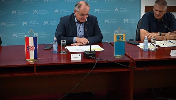 A person sitting at a table with flags of Croatia and another entity displayed on stands. - Olive Oil Times
