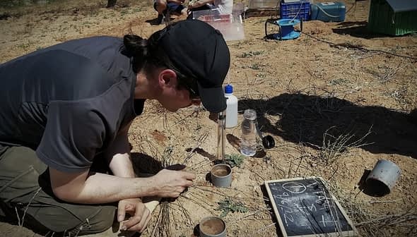 Individual wearing a cap analyzing soil samples in a field with equipment and containers nearby. - Olive Oil Times