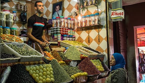 A market stall displaying a variety of olives and spices, with a vendor and a customer present. - Olive Oil Times