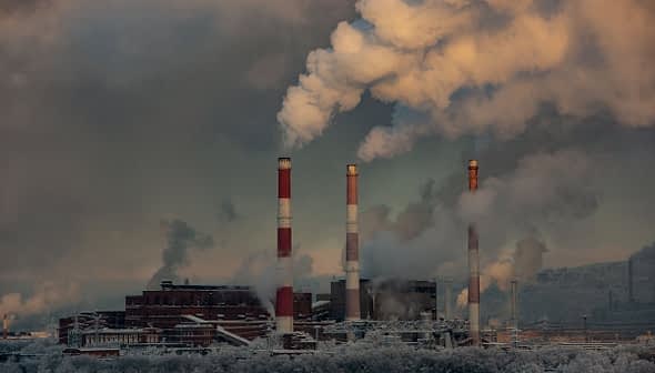 Industrial power plant with three tall smoke stacks emitting white smoke against a cloudy sky. - Olive Oil Times