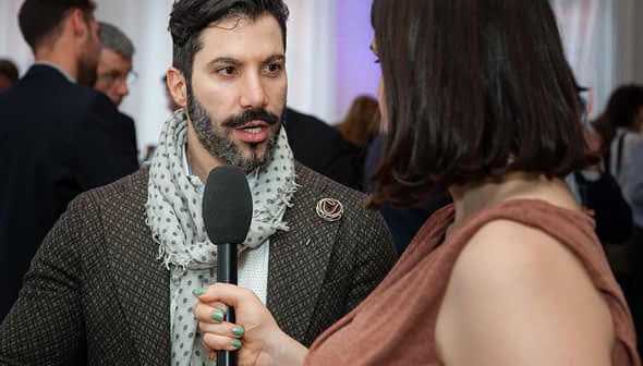 Man with a beard wearing a patterned coat and scarf speaking into a microphone during an interview. - Olive Oil Times