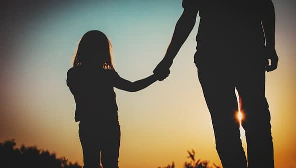 Silhouette of an adult and a child holding hands against a sunset background. - Olive Oil Times