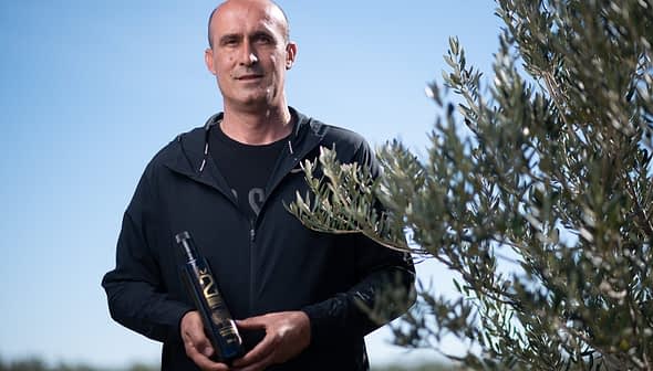 Man standing outdoors holding a bottle of olive oil next to an olive tree. - Olive Oil Times