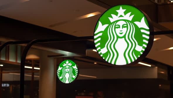 Illuminated Starbucks logo sign featuring a green background and white mermaid design. - Olive Oil Times