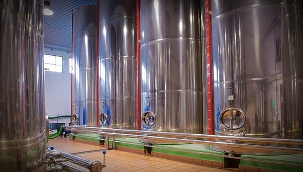 Row of stainless steel fermentation tanks used in winemaking process inside a winery. - Olive Oil Times