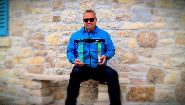 Man wearing sunglasses and a blue jacket holding two bottles of olive oil while seated on a stone bench. - Olive Oil Times