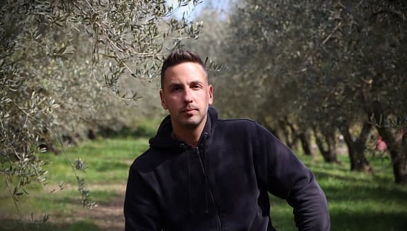 A man wearing a black hoodie sitting among olive trees in an olive grove. - Olive Oil Times