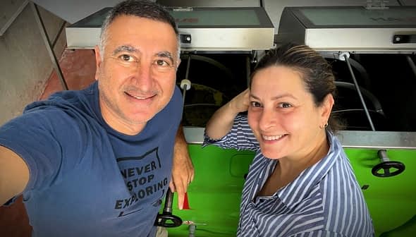 Two individuals smiling while standing next to machinery in a workshop environment. - Olive Oil Times