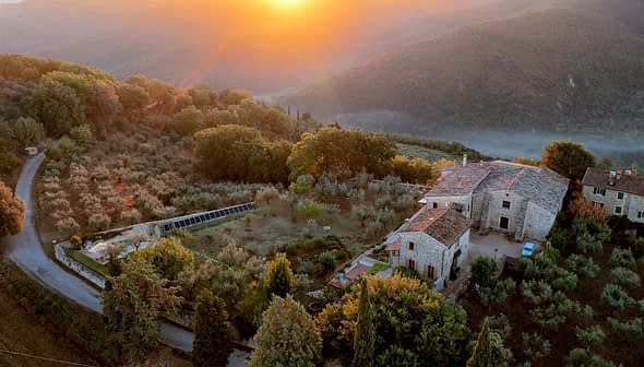 Aerial view of a rustic farmhouse surrounded by olive trees and hills during sunset. - Olive Oil Times