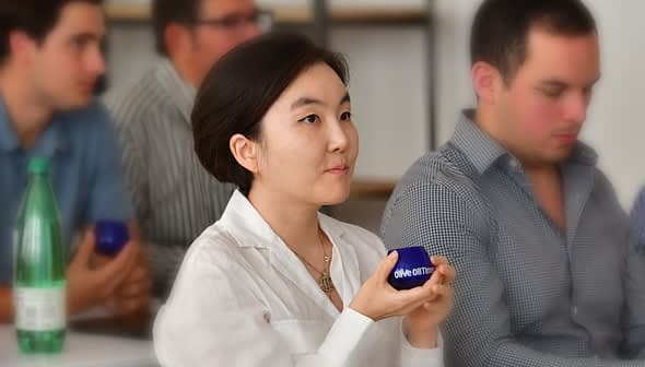 A woman with short hair holding a blue container while seated among a group of people. - Olive Oil Times