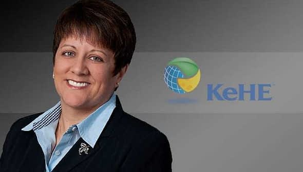 A business executive in a black suit with a light blue shirt, smiling against a dark background with the KeHE logo. - Olive Oil Times