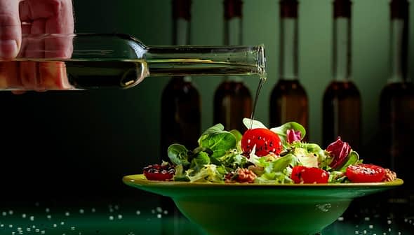 A hand pouring olive oil from a glass bottle onto a salad in a green bowl with cherry tomatoes and greens. - Olive Oil Times