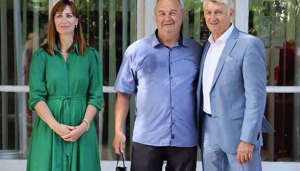 Three individuals standing together for a photo, with one woman in a green dress and two men in casual and formal attire. - Olive Oil Times