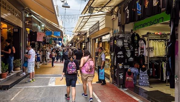 Two people walking through a market street lined with shops and stalls selling various items. - Olive Oil Times