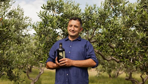 Man standing in an olive grove holding a bottle of olive oil with olive trees in the background. - Olive Oil Times
