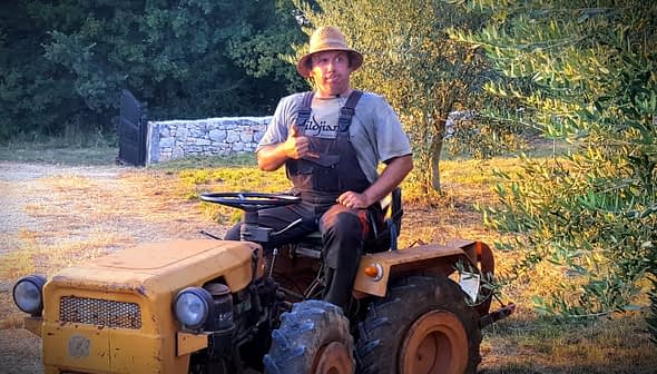 A man sitting on a yellow tractor wearing a straw hat and overalls, giving a thumbs up. - Olive Oil Times