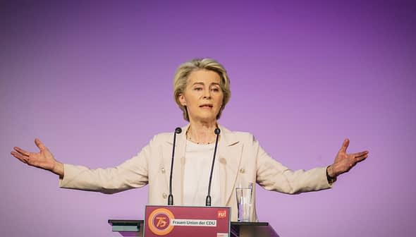 A woman gesturing while speaking at a podium during an event with a purple background. - Olive Oil Times