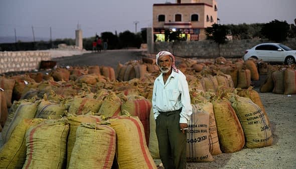 An older man wearing traditional attire stands among burlap sacks filled with an unspecified product. - Olive Oil Times