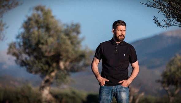 Man wearing a black polo shirt and jeans standing outdoors with olive trees in the background. - Olive Oil Times