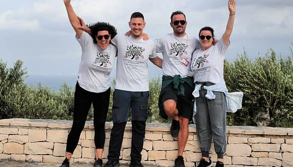 Four individuals posing together outdoors, wearing matching t-shirts and smiling at the camera. - Olive Oil Times