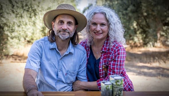 A man and woman sitting together outdoors, both smiling, with cans placed on a table in front of them. - Olive Oil Times