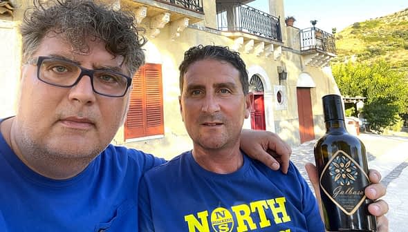 Two men posing for a selfie while holding a bottle of olive oil outdoors. - Olive Oil Times