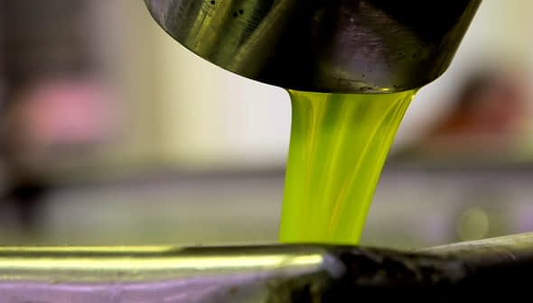 Green olive oil pouring from a metal spout into a container. - Olive Oil Times
