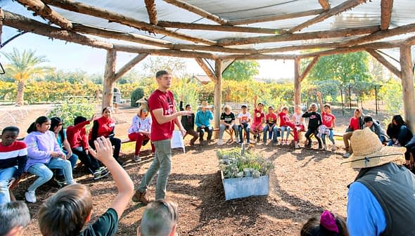 A group of children and adults seated in a circular outdoor learning space with a speaker in the center. - Olive Oil Times