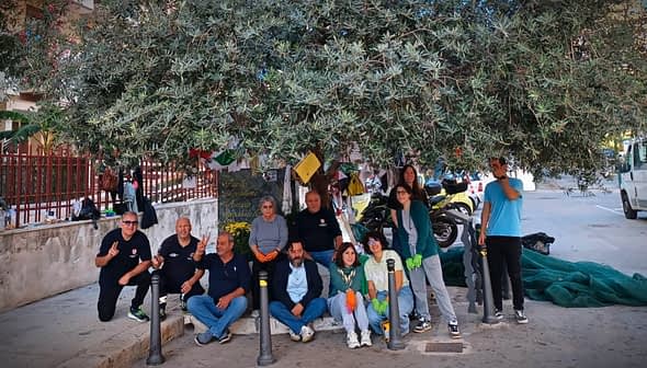 A group of people gathered under a large olive tree, some wearing gloves and smiling at the camera. - Olive Oil Times