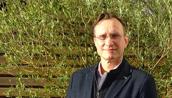 Man wearing glasses and a black jacket standing in front of green plants. - Olive Oil Times