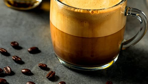 A glass mug filled with a layered coffee beverage, featuring a frothy top and dark liquid below. - Olive Oil Times