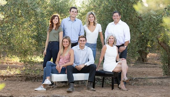 Seven individuals posing together outdoors in a natural setting with olive trees in the background. - Olive Oil Times