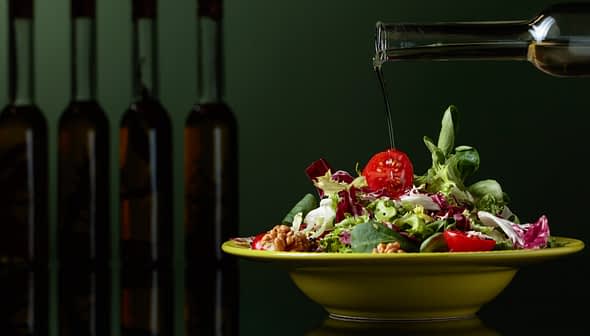 Olive oil being poured over a fresh salad in a yellow bowl with various vegetables. - Olive Oil Times