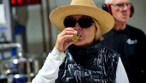 Woman wearing a straw hat and sunglasses tasting olive oil in a factory setting. - Olive Oil Times