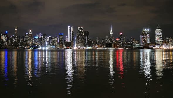 Night view of the New York City skyline with illuminated buildings reflecting on the water. - Olive Oil Times