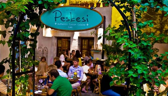 Entrance sign of Peskesi restaurant with greenery and patrons dining inside. - Olive Oil Times