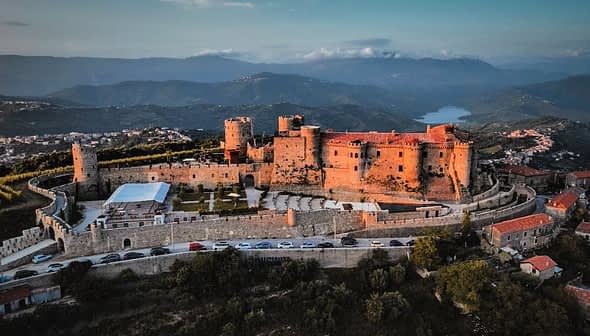 Aerial view of a historic castle surrounded by mountains and greenery. - Olive Oil Times