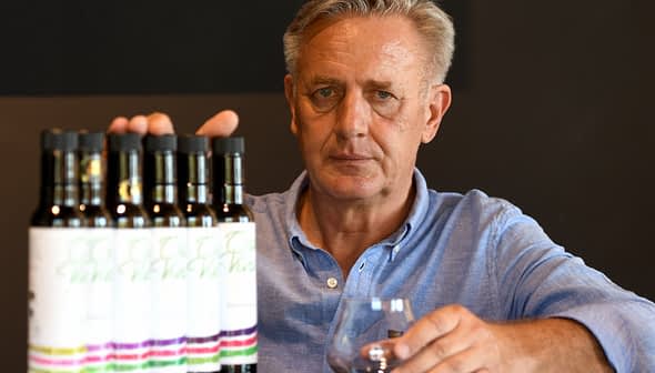Man with gray hair holding a glass in front of a row of olive oil bottles on a table. - Olive Oil Times