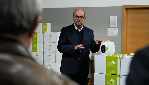 A man in a suit presenting a container of olive oil in a warehouse setting. - Olive Oil Times