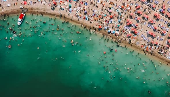 Aerial view of a crowded beach with swimmers and colorful umbrellas along the shoreline. - Olive Oil Times