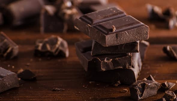 Several pieces of dark chocolate stacked on a wooden surface with additional chocolate fragments scattered around. - Olive Oil Times