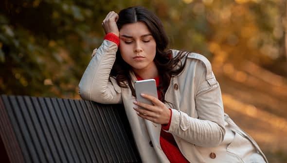 A woman with long dark hair wearing a light coat and red sweater, looking at her smartphone while sitting on a bench. - Olive Oil Times
