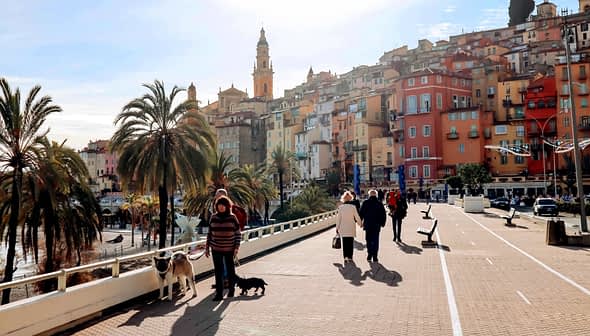 People walking along a promenade in a coastal town with colorful buildings and palm trees. - Olive Oil Times